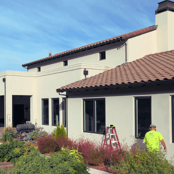 Worker in high-visibility shirt near a white Mediterranean-style home with terracotta roof and garden.
