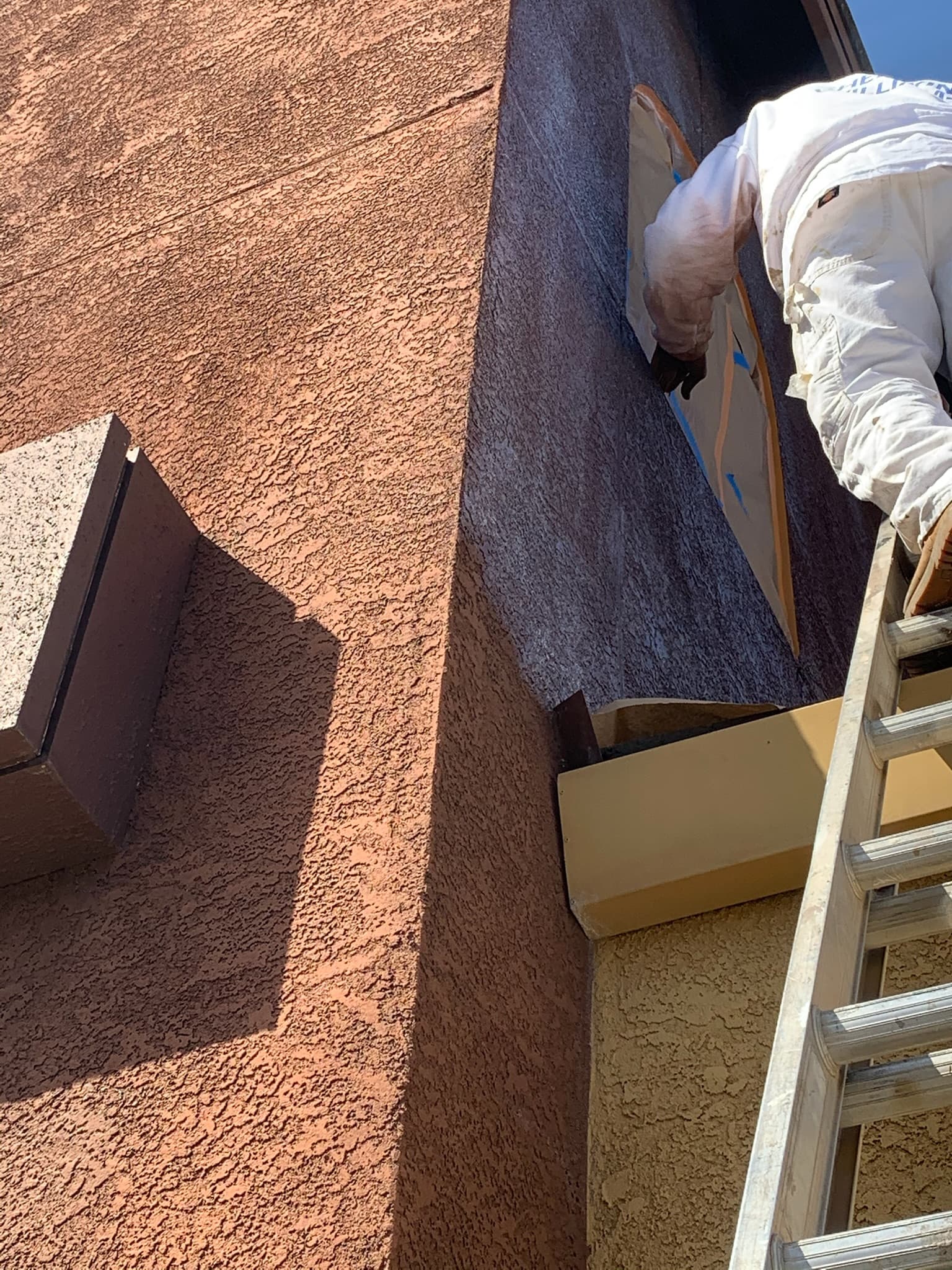 Painter in white overalls on a ladder painting a brown textured stucco exterior wall.