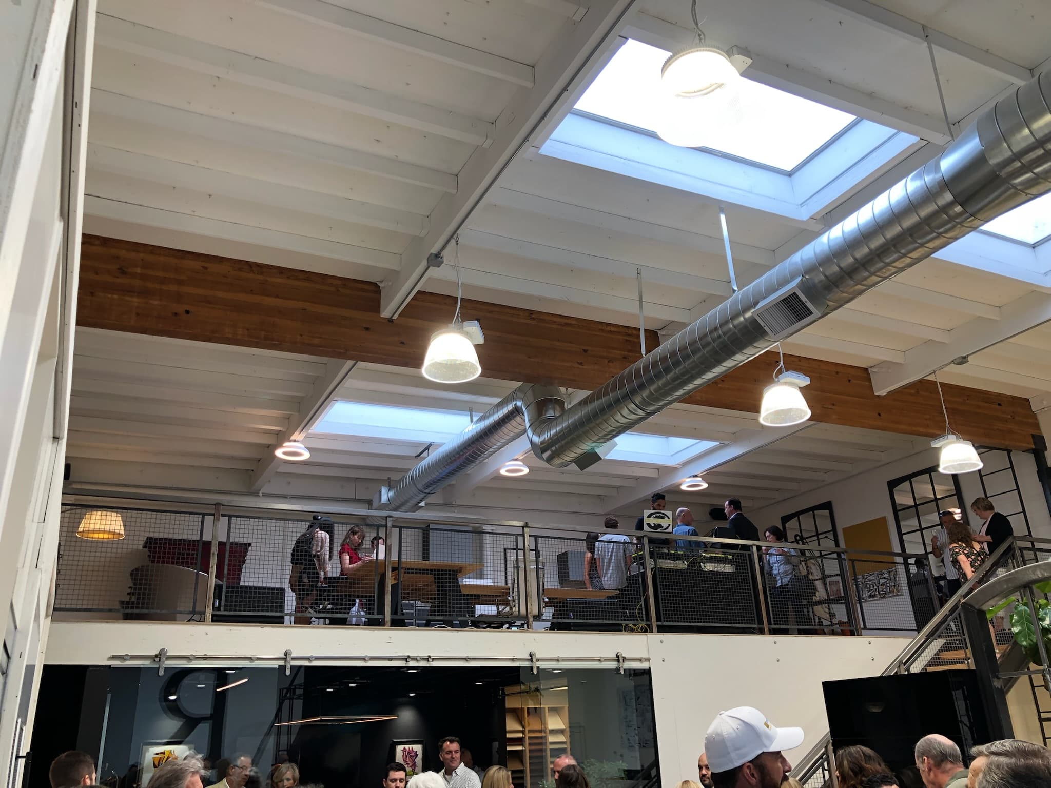People socialize in a modern building with a mezzanine, skylights, and exposed industrial ventilation.