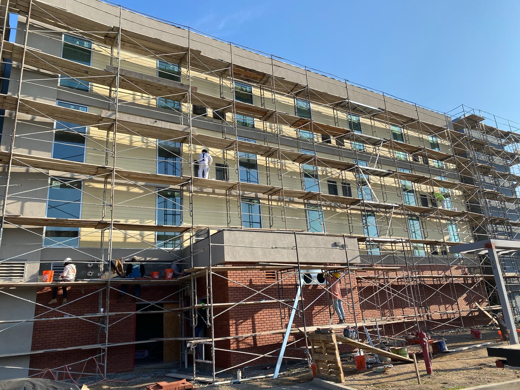 Construction workers on metal scaffolding outside a multi-story building with yellow siding and brickwork.