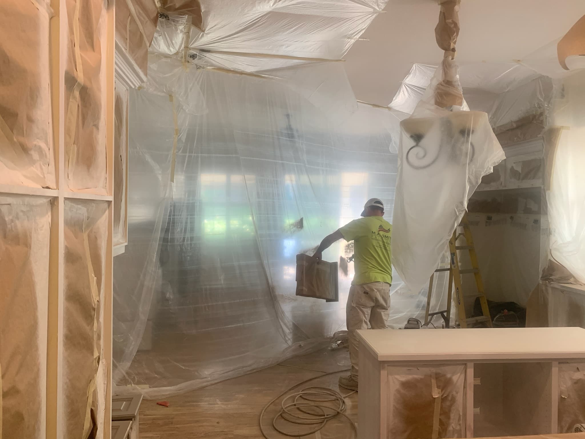 A professional painter works in a kitchen protected by extensive plastic sheeting and masking paper.