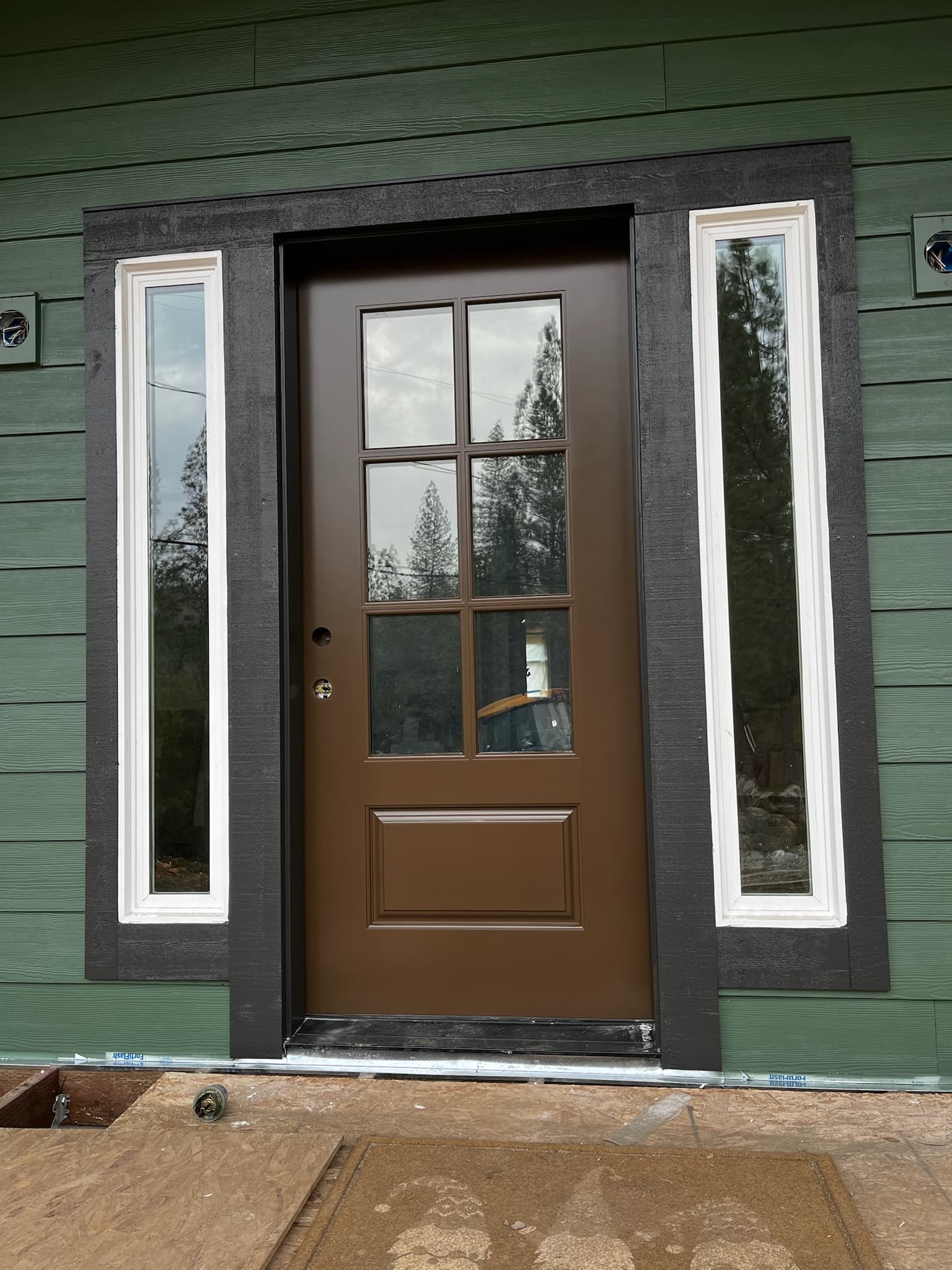 Brown front door with six glass panes and white-framed sidelights on green siding.