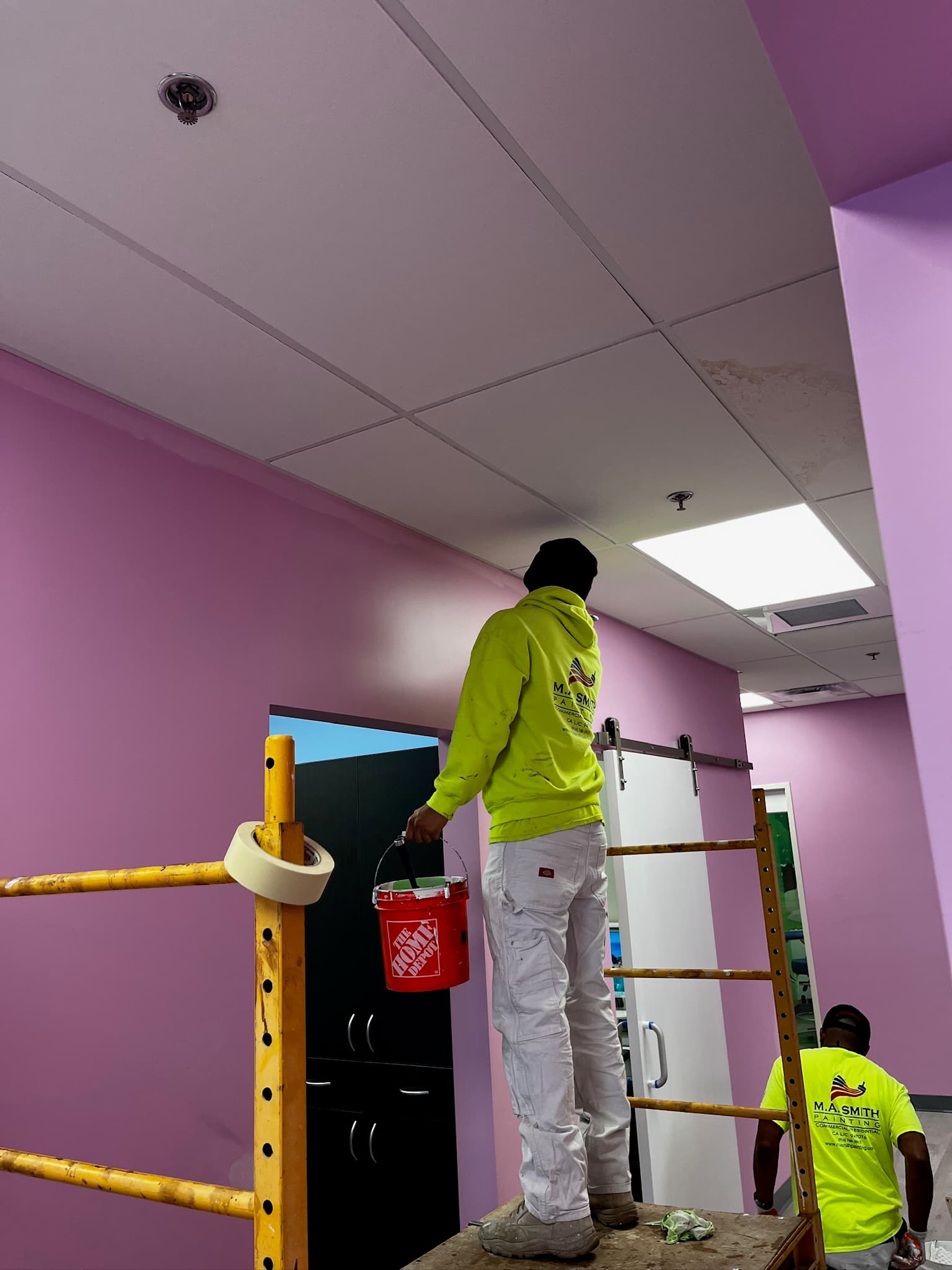 Painter in neon yellow hoodie on scaffolding holding a paint bucket against vibrant pink walls.